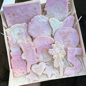 Puede incluir: Una caja de galletas de azúcar decoradas con un tema de hadas. Las galletas están decoradas con glaseado rosa y blanco y detalles dorados. Las galletas representan un hada, una casa de setas, una botella de polvo de hadas, una varita de estrella, una mariposa y las palabras "Feliz cumpleaños", "Besos de mariposa y deseos de hadas" y "Pide un deseo".
