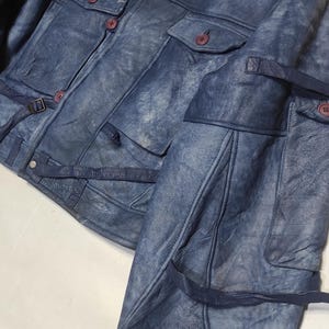 Op de afbeelding: Blau leren jas met een schapenvachtkraag. De jas heeft meerdere zakken, knoopsluitingen en verstelbare riemen. Het leer heeft een verweerde uitstraling, waardoor het een vintage look heeft.