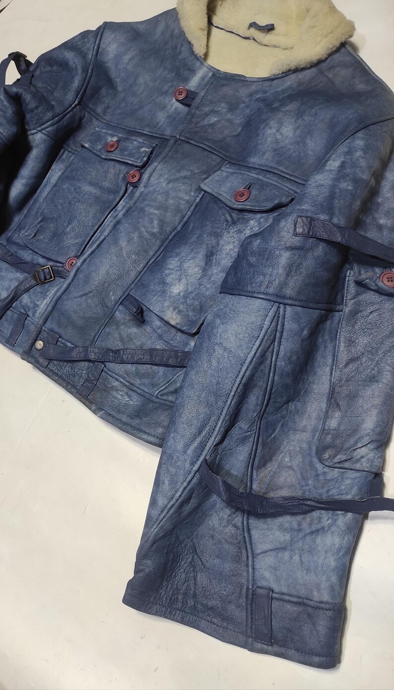 Op de afbeelding: Blau leren jas met een schapenvachtkraag. De jas heeft meerdere zakken, knoopsluitingen en verstelbare riemen. Het leer heeft een verweerde uitstraling, waardoor het een vintage look heeft.