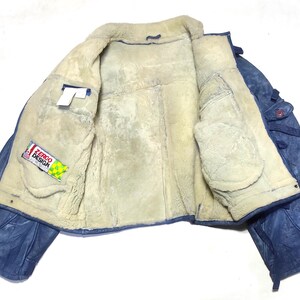 Op de afbeelding: Open weergave van een blauwe leren jas met een cr&egrave;mekleurige shearling voering. De jas heeft twee voorzakken en een label met de tekst "ZERGO DESIGN". De jas ligt plat en toont zijn binnenkant.