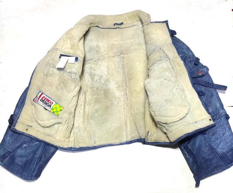 Op de afbeelding: Open weergave van een blauwe leren jas met een cr&egrave;mekleurige shearling voering. De jas heeft twee voorzakken en een label met de tekst "ZERGO DESIGN". De jas ligt plat en toont zijn binnenkant.