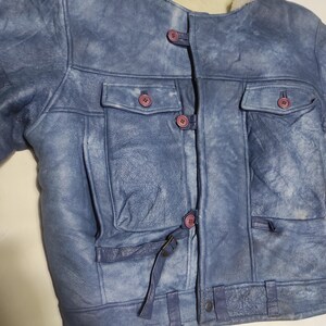 Op de afbeelding: Blau leren jas met een cr&egrave;me kleurige schapenvacht kraag. De jas heeft twee borstzakken, twee zijzakken en een knoopsluiting met roze knopen. De jas heeft een verweerde uitstraling.