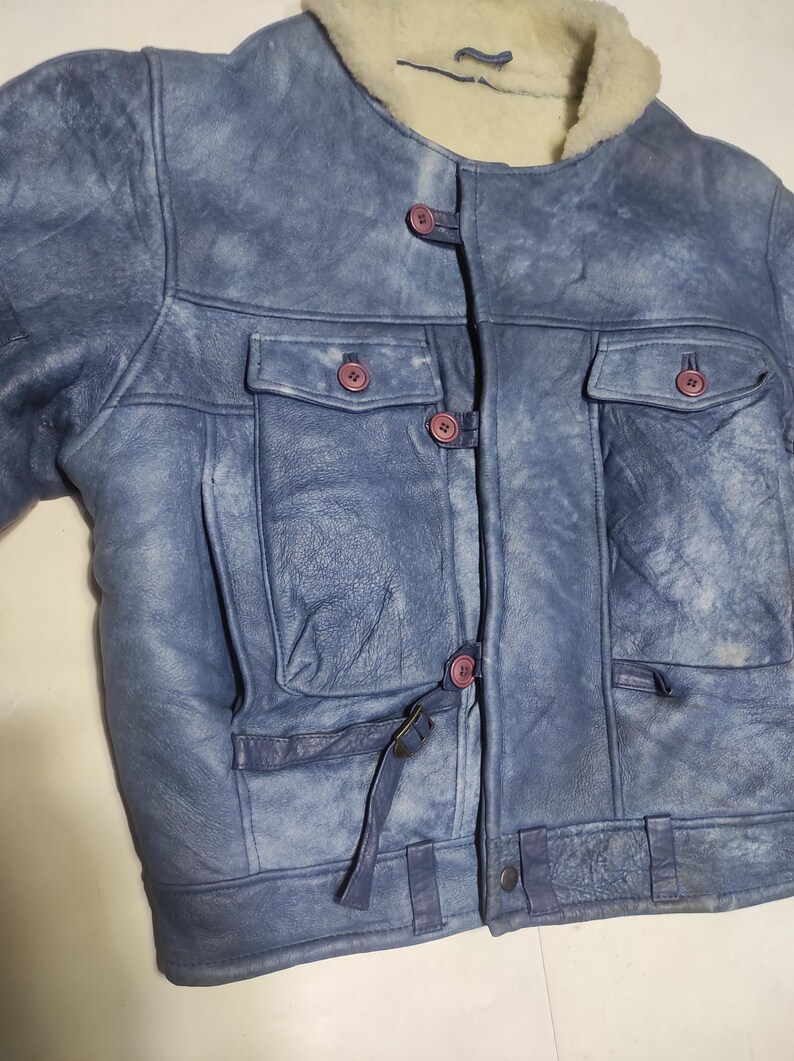 Op de afbeelding: Blau leren jas met een cr&egrave;me kleurige schapenvacht kraag. De jas heeft twee borstzakken, twee zijzakken en een knoopsluiting met roze knopen. De jas heeft een verweerde uitstraling.