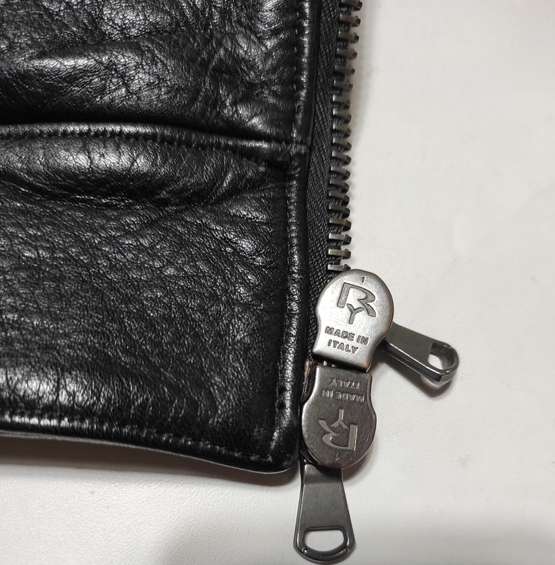 May include: Close-up of a black leather jacket zipper with a silver zipper pull that says "Made in Italy" and has the letter "R" on it.
