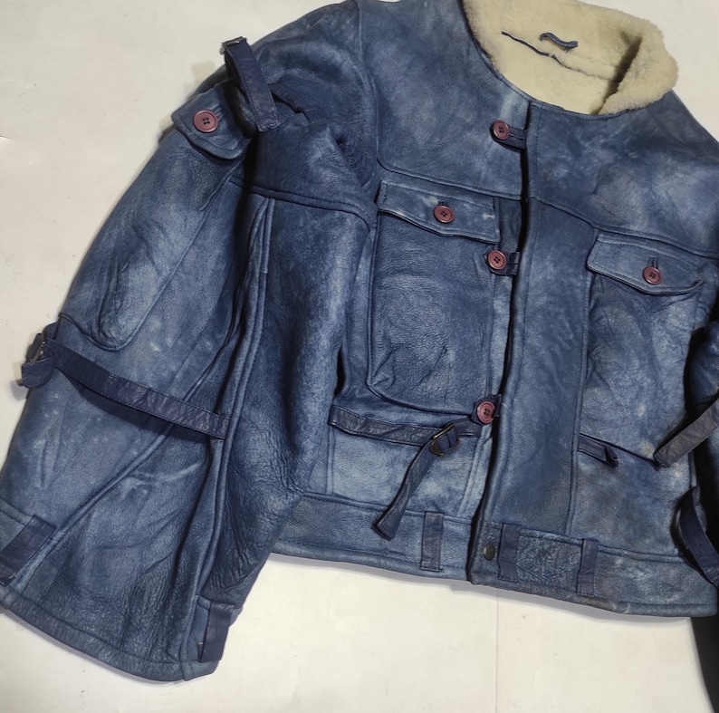 Op de afbeelding: Blau leren jas met een cr&egrave;me kleurige schapenvacht kraag. De jas heeft meerdere zakken, knopen en bandjes. Het leer heeft een verweerde uitstraling en de knopen zijn roodbruin. De jas is in vintage stijl.