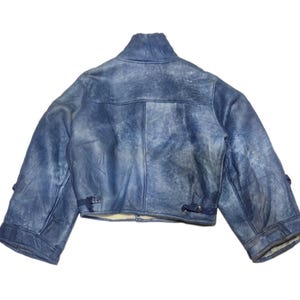 Op de afbeelding: Een korte, blauwe spijkerjas met een hoge kraag. De jas heeft een verweerde, vervaagde uitstraling en heeft twee kleine gespen op elke mouw. De binnenvoering is licht van kleur.