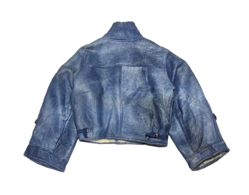 Op de afbeelding: Een korte, blauwe spijkerjas met een hoge kraag. De jas heeft een verweerde, vervaagde uitstraling en heeft twee kleine gespen op elke mouw. De binnenvoering is licht van kleur.