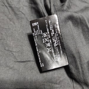 May include: Black care label with white text. The label includes the style and model number, size, fabric composition, care instructions, and country of origin. The text reads: "Style/Model LET013 WOF01 Fabric Shell: COW Lining: polyester 60% cupra 40% cotton 95% polyurethane 5% Made in Japan"