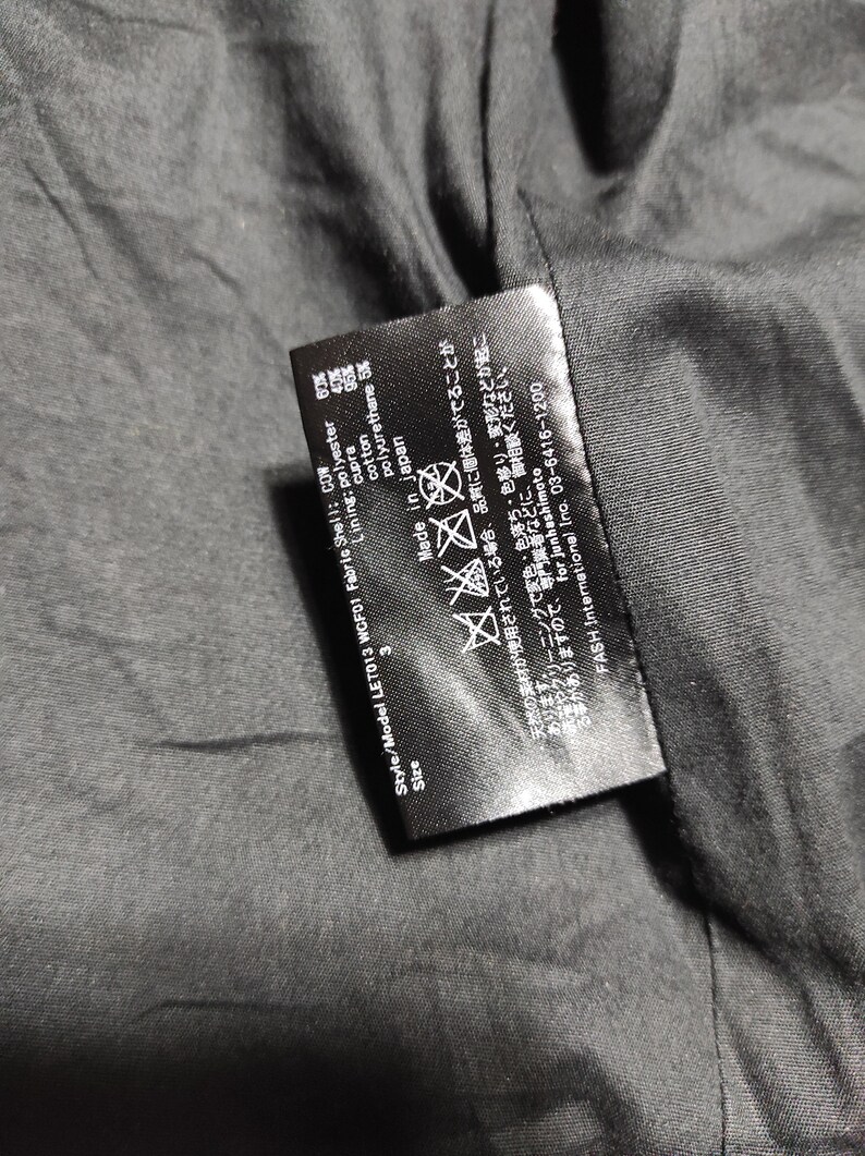 May include: Black care label with white text. The label includes the style and model number, size, fabric composition, care instructions, and country of origin. The text reads: "Style/Model LET013 WOF01 Fabric Shell: COW Lining: polyester 60% cupra 40% cotton 95% polyurethane 5% Made in Japan"