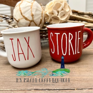 May include: Two white ceramic mugs with red handles. One mug says "Jax" in red lettering and the other mug says "Storie" in white lettering.