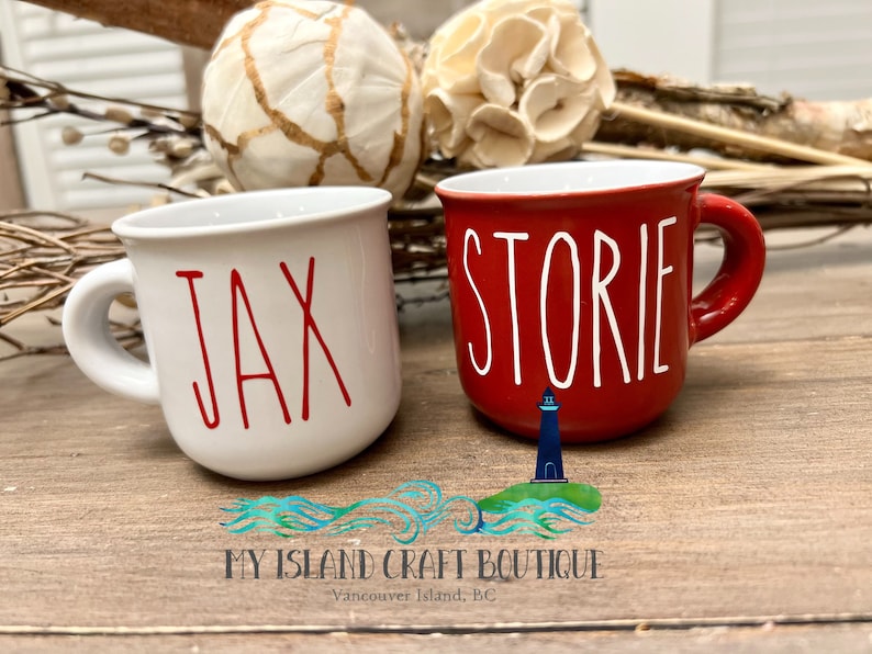 May include: Two ceramic mugs with rounded handles. The white mug features the red text JAX, while the red mug features the white text STORIE. Both mugs have a smooth, glossy finish and a simple, functional design for holding hot beverages.
