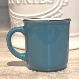 May include: A solid teal ceramic mug with a smooth finish and a dark rim along the top edge. The mug features a curved handle on the left side and a white interior. The design is simple and functional, with a rounded base and a slightly flared rim.