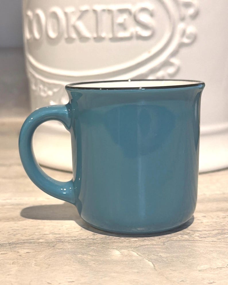 May include: A solid teal ceramic mug with a smooth finish and a dark rim along the top edge. The mug features a curved handle on the left side and a white interior. The design is simple and functional, with a rounded base and a slightly flared rim.