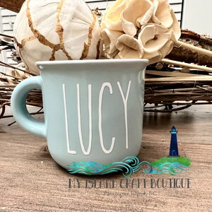 May include: A light blue ceramic mug featuring the name LUCY written in white, tall, thin, sans-serif capital letters. The mug has a rounded body and a curved handle on the left side, presenting a smooth, solid-colored finish throughout the exterior.