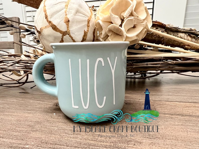 May include: A light blue ceramic mug featuring the name LUCY written in white, tall, thin, sans-serif capital letters. The mug has a rounded body and a curved handle on the left side, presenting a smooth, solid-colored finish throughout the exterior.