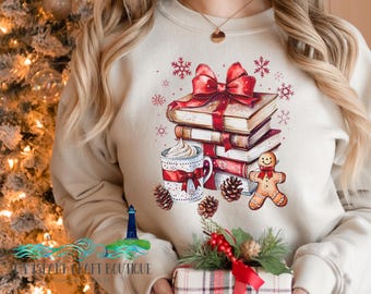 May include: A cream-coloured crewneck sweatshirt featuring a graphic print of a stack of books topped with a large red bow, a mug of hot cocoa with whipped cream, pinecones, and a gingerbread cookie, surrounded by small red snowflake illustrations.