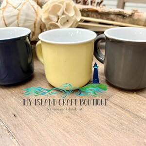 May include: Three ceramic mugs in navy blue, pale yellow, and charcoal gray, each featuring a white interior and a curved handle. A logo with a lighthouse icon and the text MY ISLAND CRAFT BOUTIQUE Vancouver Island, BC is centered below.