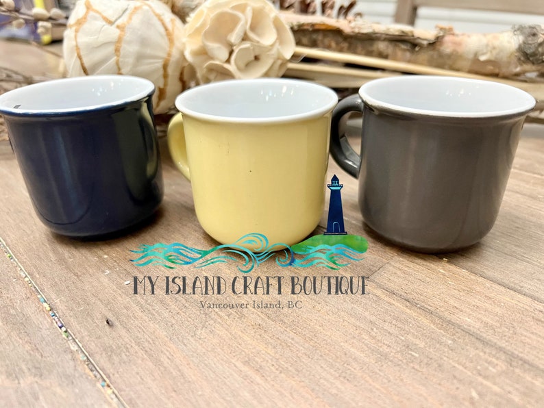 May include: Three ceramic mugs in navy blue, pale yellow, and charcoal gray, each featuring a white interior and a curved handle. A logo with a lighthouse icon and the text MY ISLAND CRAFT BOUTIQUE Vancouver Island, BC is centered below.
