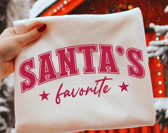May include: A white sweatshirt with the text "SANTA'S favorite" in red script. The sweatshirt is held by a hand with red nail polish. The background includes a blurred Christmas scene with lights and snow.