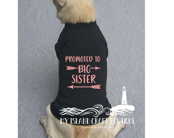 May include: A black dog shirt with the text "Promoted to Big Sister" in pink with arrows on either side. The shirt is on a small dog with a light brown coat. The text "My Island Craft Boutique" is at the bottom of the shirt.