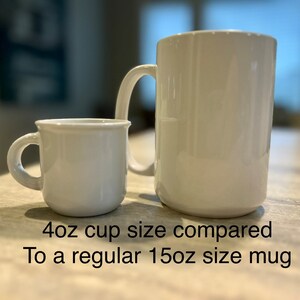 May include: A small white 4oz cup positioned next to a larger white 15oz mug. The text below the items reads: 4oz cup size compared To a regular 15oz size mug. Both items feature a smooth, glossy finish and a single handle on the side.