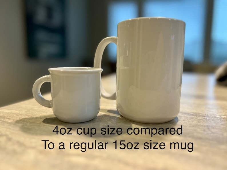 May include: A small white 4oz cup positioned next to a larger white 15oz mug. The text below the items reads: 4oz cup size compared To a regular 15oz size mug. Both items feature a smooth, glossy finish and a single handle on the side.