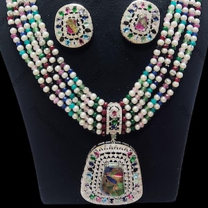 Könnte beinhalten: Ein mehrreihiges Perlenketten- und Ohrring-Set. Die Halskette besteht aus Strängen aus weißen Perlen, die mit farbigen Perlen durchsetzt sind. Anhänger und Ohrringe haben eine silberfarbene Fassung mit bunten Edelsteinen.
