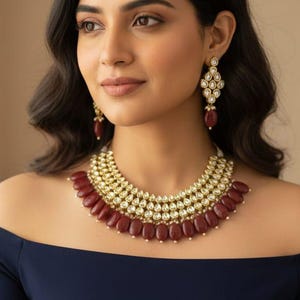 May include: A gold-toned necklace and matching earrings set with ruby-colored stones. The necklace features multiple layers of small, round gold accents and larger, oval ruby stones. The earrings have a similar design, with ruby drops.