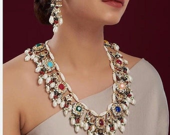 Op de afbeelding: Een gouden ketting met een veelkleurig stenen ontwerp en parel accenten. De ketting is gelaagd met kleine witte parels en heeft een verscheidenheid aan gekleurde stenen, waaronder rood, blauw, groen en roze. De ketting wordt gedragen door een persoon met donker haar en een lichtgekleurde top.