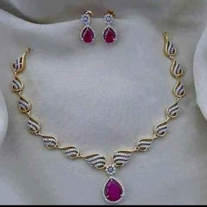 May include: A gold necklace with a teardrop-shaped pendant set with a large red gemstone. The necklace is adorned with smaller gemstones and has a delicate, intricate design. Matching earrings with red gemstones are also included.