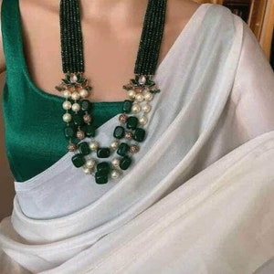May include: A white and green beaded necklace with a floral design. The necklace is made of green and white beads and has a floral design in the center. The necklace is worn by a person wearing a green blouse and a white saree.