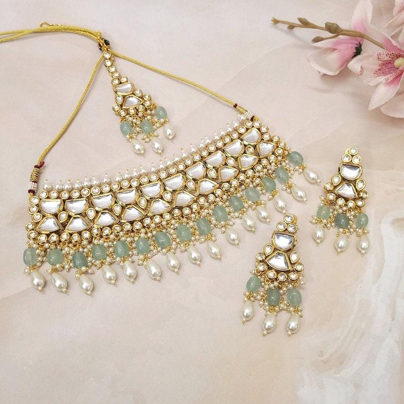 Puede incluir: Un juego de collar de oro con un dise&ntilde;o de piedras blancas y verdes. El collar tiene un estilo choker con un gran colgante y pendientes a juego. Las piedras est&aacute;n engastadas en un patr&oacute;n geom&eacute;trico y el collar est&aacute; adornado con perlas.