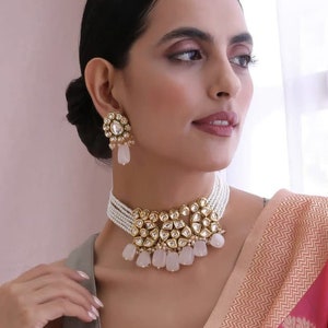 May include: A white pearl choker necklace with a gold and white stone pendant. The pendant features a floral design with a teardrop-shaped center stone. The necklace is accented with white stone drops.