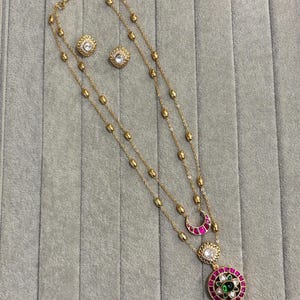 Può includere: Una collana d'oro con una mezzaluna e un ciondolo rotondo con pietre verdi e rosse. La collana è a strati e viene fornita con un paio di orecchini abbinati con pietre trasparenti.