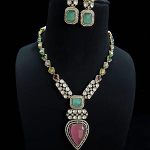 May include: A gold necklace featuring a pink teardrop-shaped pendant and a green square pendant. The necklace is decorated with clear and colourful stones. Matching earrings with green square stones are also included.