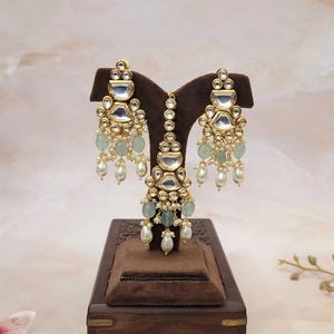 Puede incluir: Un conjunto de pendientes y maang tikka de tono dorado con un dise&ntilde;o de l&aacute;grima. El conjunto presenta piedras transparentes y verdes, y perlas blancas.