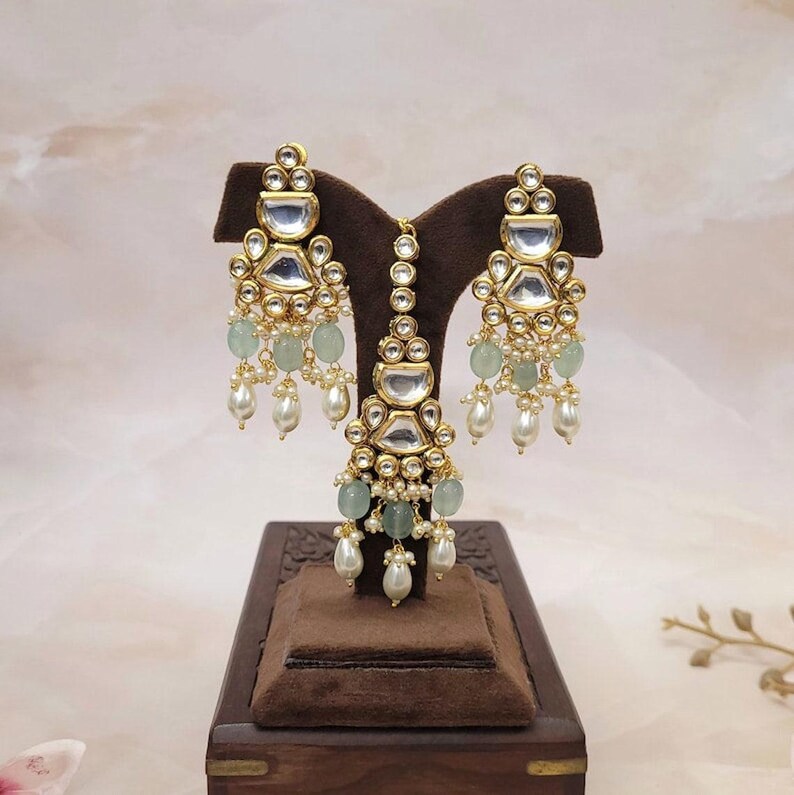 Puede incluir: Un conjunto de pendientes y maang tikka de tono dorado con un dise&ntilde;o de l&aacute;grima. El conjunto presenta piedras transparentes y verdes, y perlas blancas.