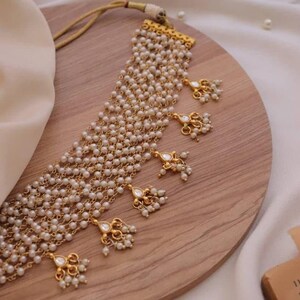 Puede incluir: Un collar de oro con m&uacute;ltiples hileras de perlas blancas y detalles dorados. El collar tiene varios colgantes con detalles de perlas.