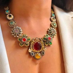 Peut inclure: Un collier en or avec un grand pendentif en pierre rouge entouré de pierres précieuses plus petites de différentes couleurs, notamment le vert, le bleu et le jaune. Le collier est en métal et présente un motif floral.