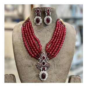 May include: A red beaded necklace with a large, ornate pendant featuring a red gemstone. The pendant is surrounded by smaller, clear gemstones. The necklace is paired with matching earrings with red gemstones.