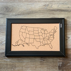 May include: A framed corkboard featuring a laser-cut outline of the United States map. The black frame surrounds the tan cork, with state borders clearly defined. The frame has a rectangular shape and is suitable for pinning notes or displaying travel memories.
