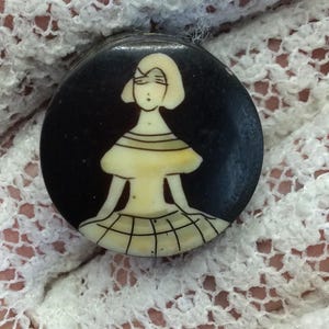 Puede incluir: Un botón negro circular con una ilustración gráfica de color crema de una figura con corte de pelo bob, que lleva una blusa de manga corta y una falda con patrón de cuadrícula. El botón tiene un acabado oscuro, liso y perfil plano.