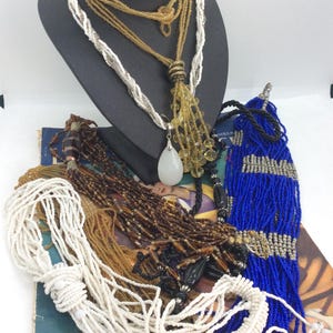 Puede incluir: Una colección de collares de cuentas en varios estilos y colores. Los collares incluyen cuentas blancas, doradas, marrones y azules. Algunos collares se exhiben en una cabeza de maniquí negra, mientras que otros están sobre un libro.