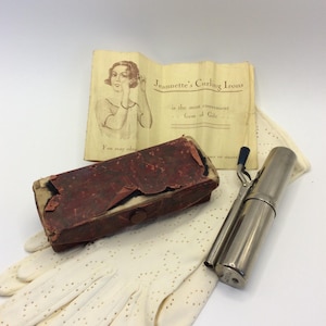 May include: A vintage metal curling iron with a black handle, a worn rectangular red box, and a pair of white perforated gloves. An off-white paper insert features an illustration of a person using the tool and the text Jeannette's Curling Irons.