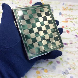 May include: A rectangular metal cigarette case featuring a polished silver and dark green checkered pattern on the front surface. The case has a smooth, reflective finish with a grid of alternating metallic silver and deep green squares.