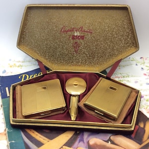 May include: A gold-tone KIGU Compacts of Character set in a presentation box. The set includes two square, textured gold-tone compacts and a round gold-tone lipstick holder, all nestled in a dark red satin-lined interior with gold lid.