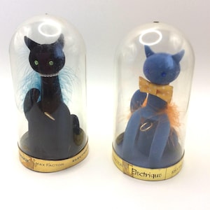 Könnte beinhalten: Zwei Max Factor Electrique Katzenfiguren in durchsichtigen Kunststoffkuppeln. Die schwarze Katze hat grüne Augen, ein perlenartiges Halsband und blaue Federn. Die blaue Katze trägt eine gelbe Fliege mit Punkten und orangefarbene Federn am Sockel.