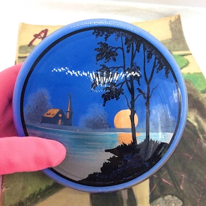 Puede incluir: Un plato decorativo redondo azul con una escena de paisaje. La imagen representa una iglesia, árboles y una puesta de sol sobre el agua. El borde del plato es azul claro y la obra de arte está detallada con detalles en negro y naranja.