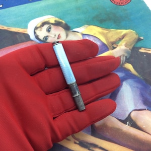 Op de afbeelding: Een vintage sigarettenhouder met een lichtblauw en zilver ontwerp. De houder wordt vastgehouden in een rode handschoen, met een vintage magazinecover op de achtergrond. De magazinecover toont de woorden "Dream of Romance".