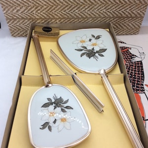 Könnte beinhalten: Ein Vintage-Kosmetikset mit einem Handspiegel, einer Bürste und einem Kamm, alle mit goldfarbenen Griffen und weißen Blumenmustern. Das Set wird in einer Box mit gewebter Textur präsentiert.